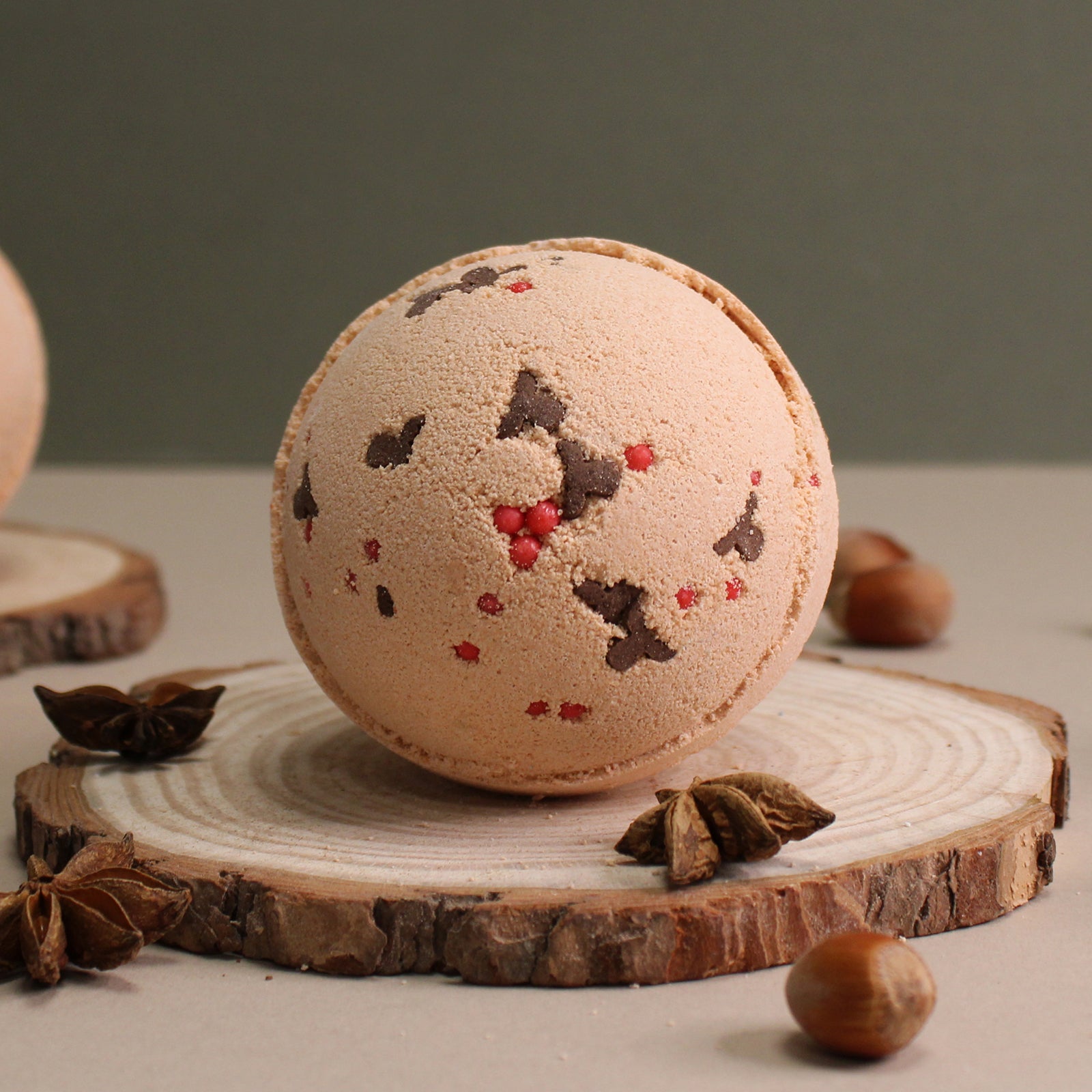 The Toffee and Caramel Reindeer 3D Bath Bomb from Scented Aromas, with its round textured design and chocolate and red detailing, is elegantly displayed on a sliced wood piece alongside a sprinkle of nuts and star anise, offering an inviting aroma.