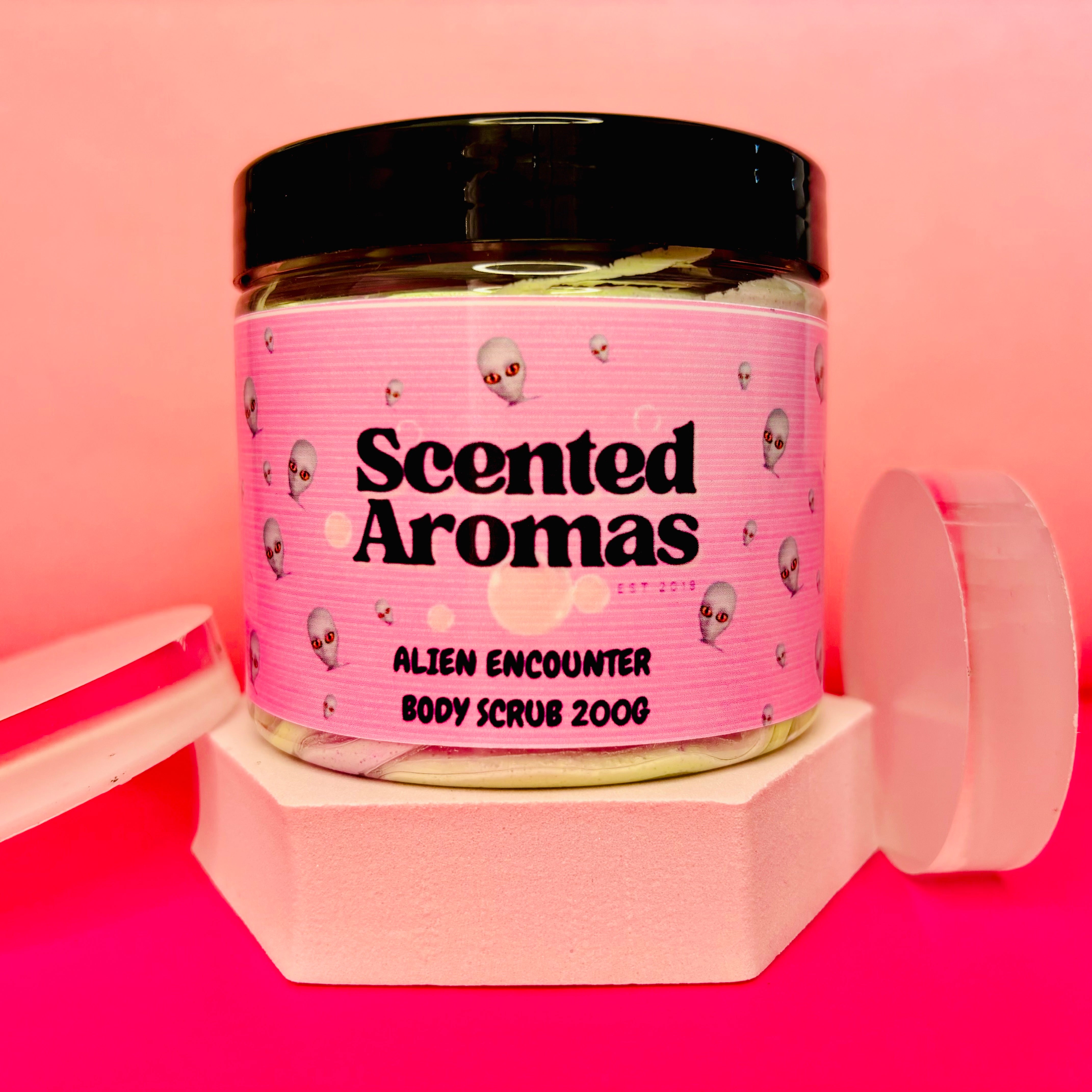 A 200g jar of Scented Aromas Alien Encounter Sugar Body Scrub sits on a hexagonal stand against a pink backdrop. Its pink label with alien head illustrations invites you to indulge in its extraterrestrial fragrance.