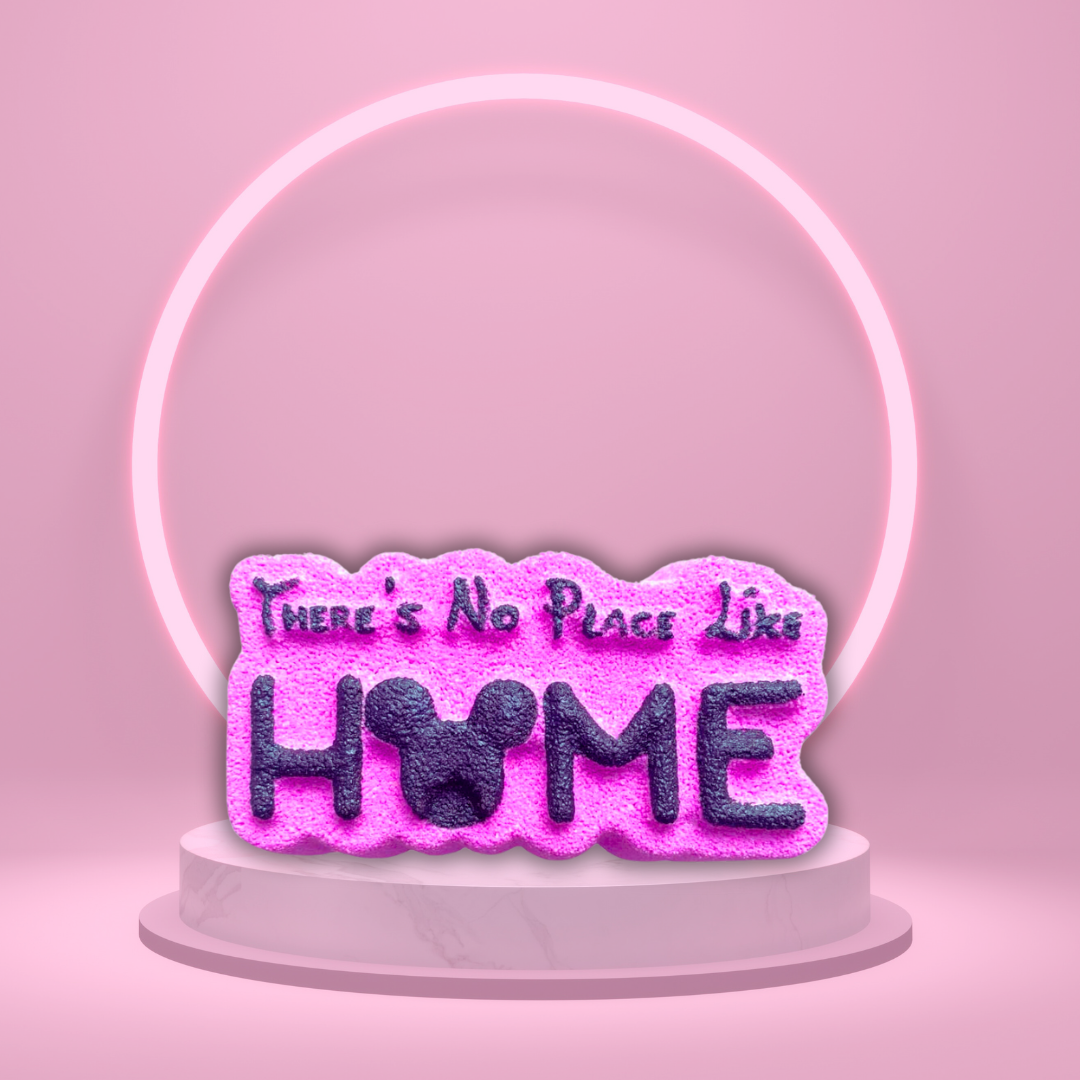 A pink neon sign with the text "There Is Only One Place Like Home," featuring a mouse-shaped "O," stands on a white pedestal against a pink background with a circular light behind it. This enchanting display evokes the magical land of Oz, adding a whimsical touch to your Magical Skincare and Home Fragrance range from The Soap Gal x.