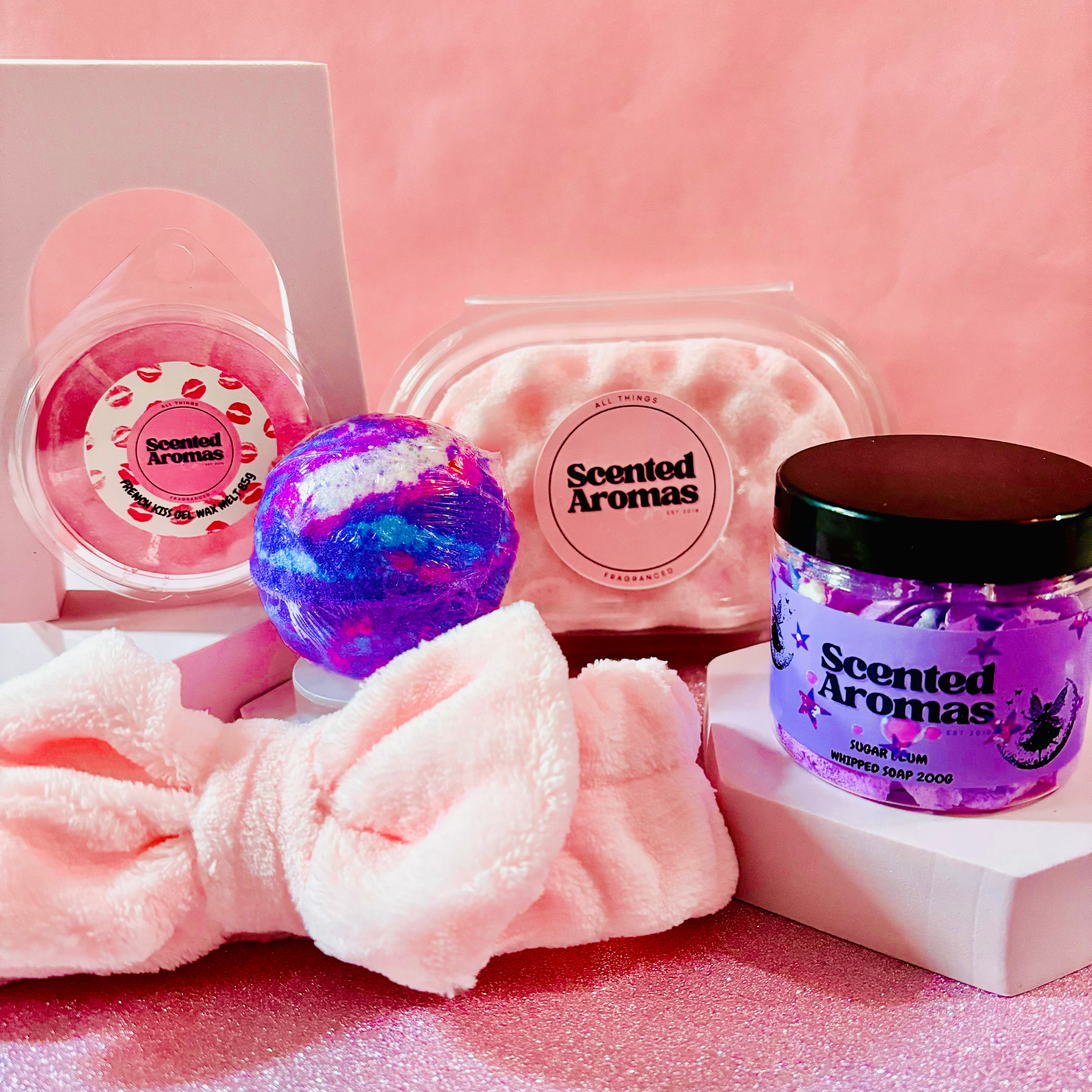 The Valentines Gift Set Bundle by Scented Aromas includes a Birthday Cake Bath Bomb, scrub, and plush headband on a glittery surface with a pink backdrop. Ideal for pamper nights or as a thoughtful gift.