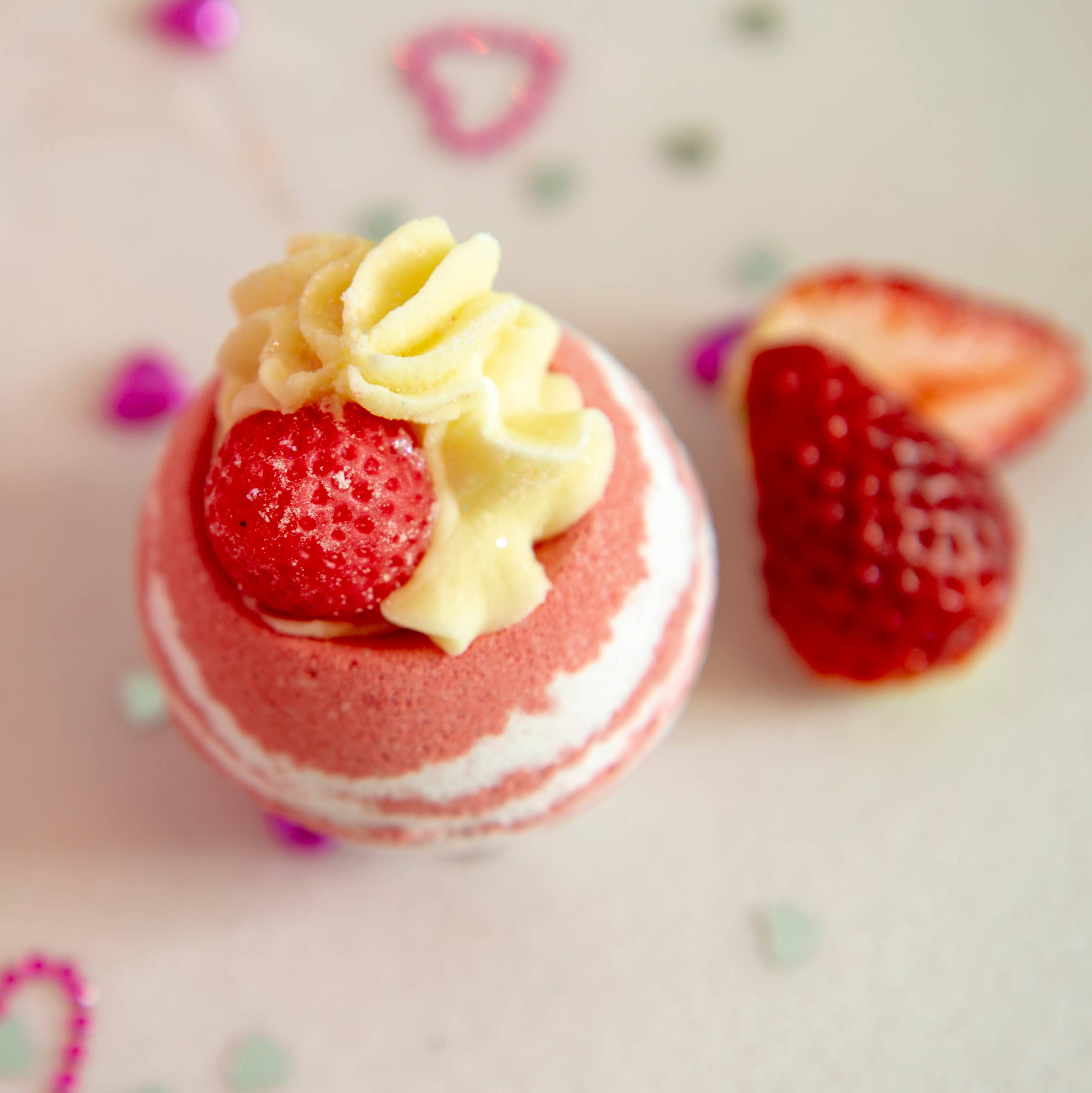 A Strawberry Prosecco Cocoa Shea Butter Bath Bomb by Scented Aromas, in pink and white hues, is positioned on a white surface surrounded by heart-shaped decorations and strawberry halves. The setting glimmers with bio glitter, infusing an enchanting sparkle into the delightful arrangement.