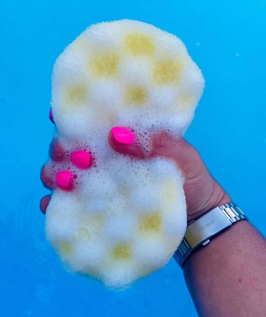 A hand with pink nail polish and a silver watch holds a soapy yellow Citronella Soap Sponge - Natural Insect Repellent from The Soap Gals against a blue background.