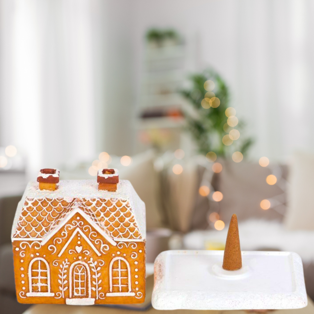 A Gingerbread House Incense Cone Burner by The Soap Gal x, featuring a removable roof and a delightful cone inside, serves as charming holiday decor. The background is a softly blurred living room adorned with string lights and complemented by a potted plant, creating a soothing atmosphere.