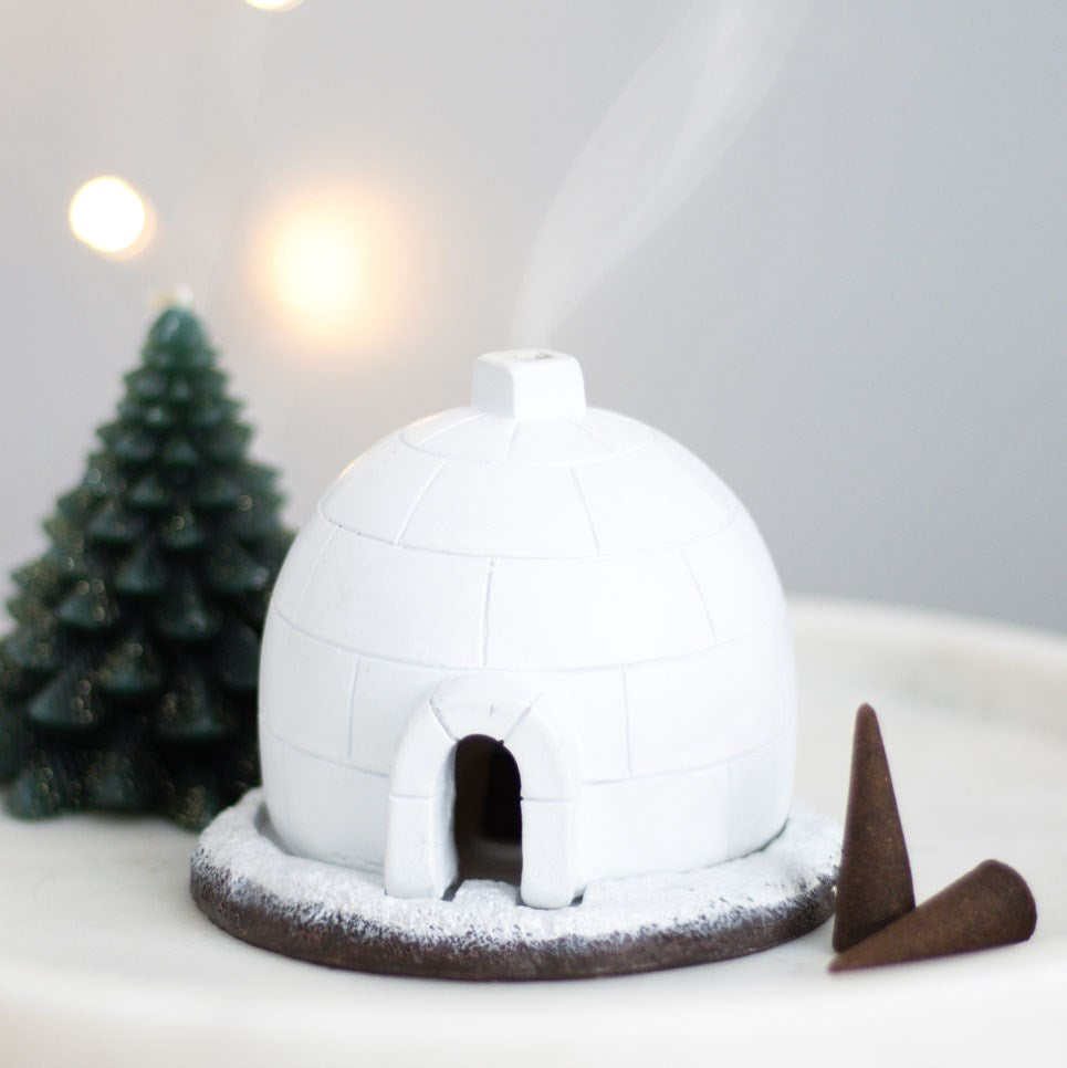 An Igloo Incense Burner by The Soap Gal x is positioned on a white surface, emitting steam that creates a holistic atmosphere. Nearby, a miniature tree and cone-shaped objects contribute to the winter-themed decor.